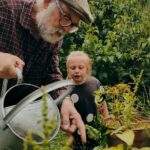 Older man watering garden with watering can, young girl watching and learning