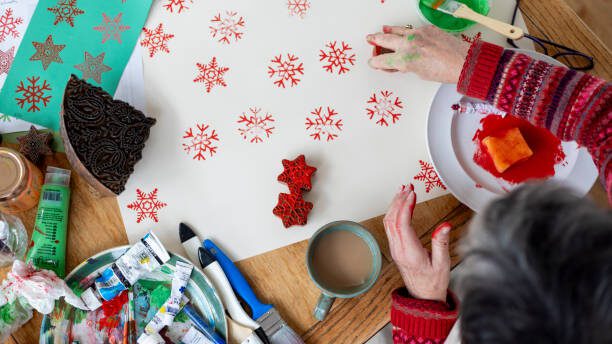An aerial shot of an unrecognisable female's hands creating patterns with a wooden carved printing block. She is sitting in the kitchen of her home in Hexham, Northumberland, enjoying a craft session. On the table there are large pieces of printed paper alongside art and crafts materials.