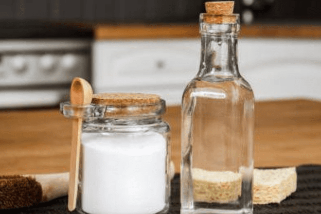 Bottle of vinegar and jar of baking soda on kitchen bench with oven in background.